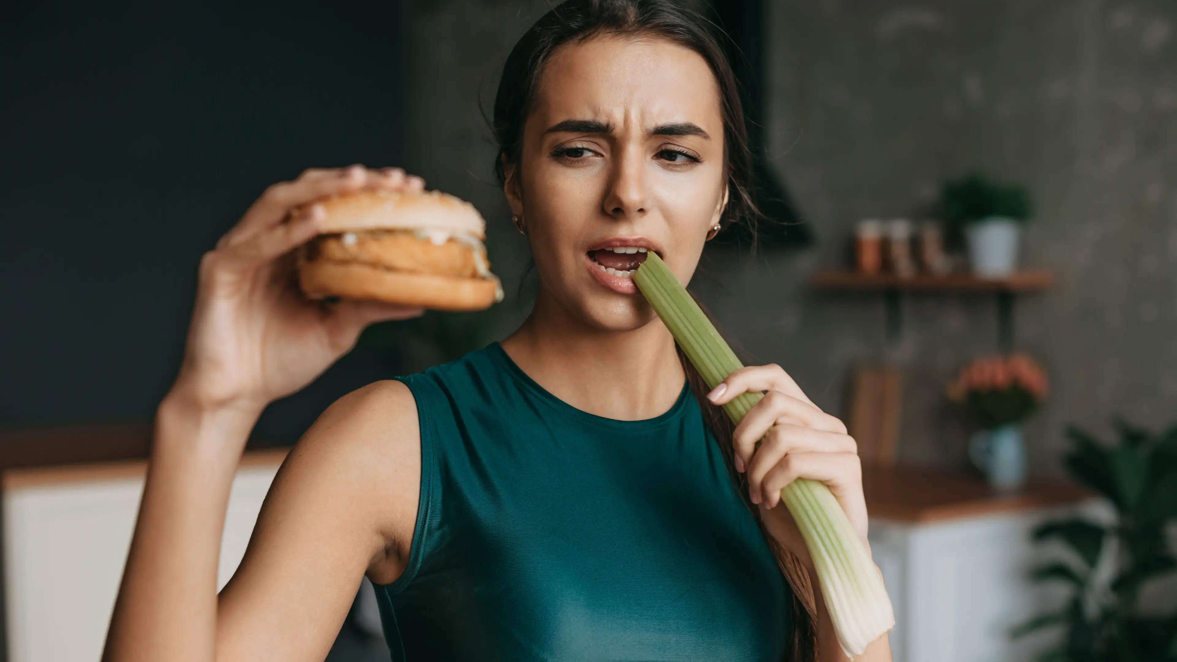 Eine Frau beißt in eine Stange Sellerie und schaut auf einen Burger in ihrer anderen Hand