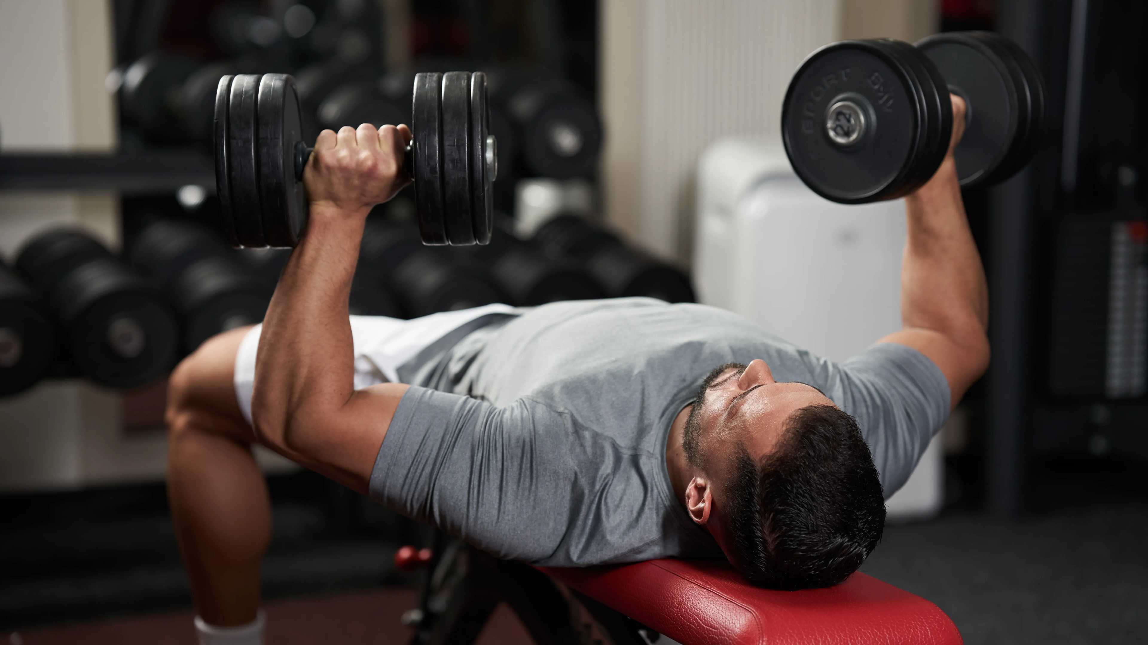 Ein Mann im T-Shirt beim Flachbankdrücken mit Kurzhanteln im Fitnessstudio