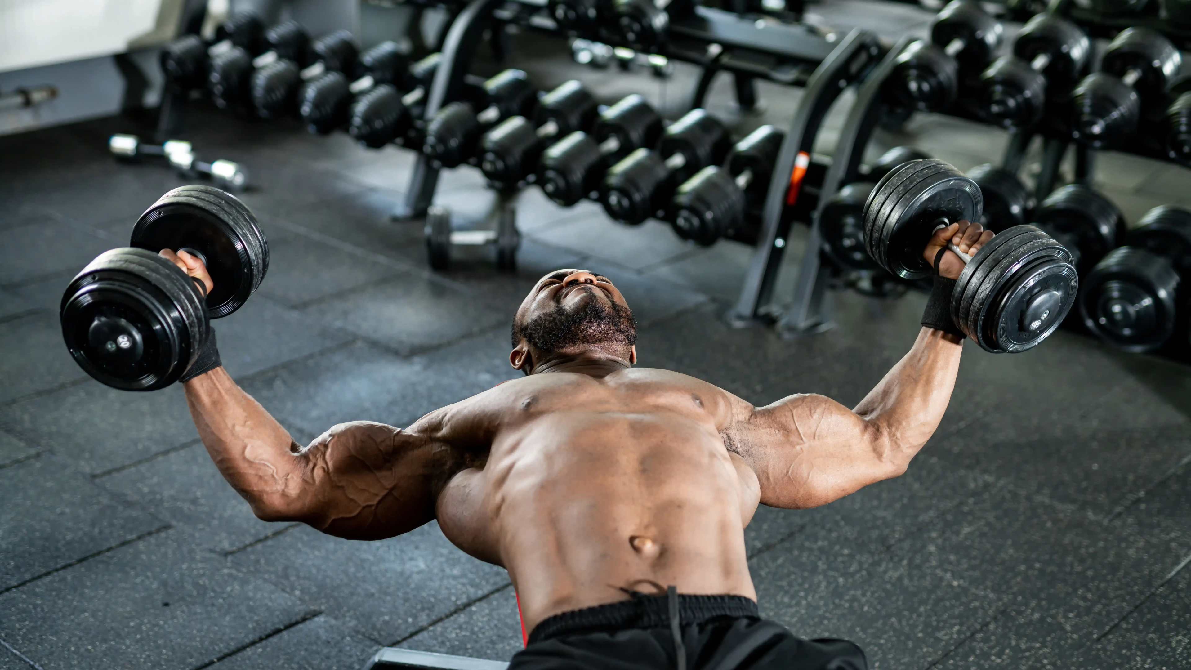 Ein oberkörperfreier, muskulöser Mann macht Chest Flys mit Kurzhanteln auf einer Flachbank im Fitnessstudio