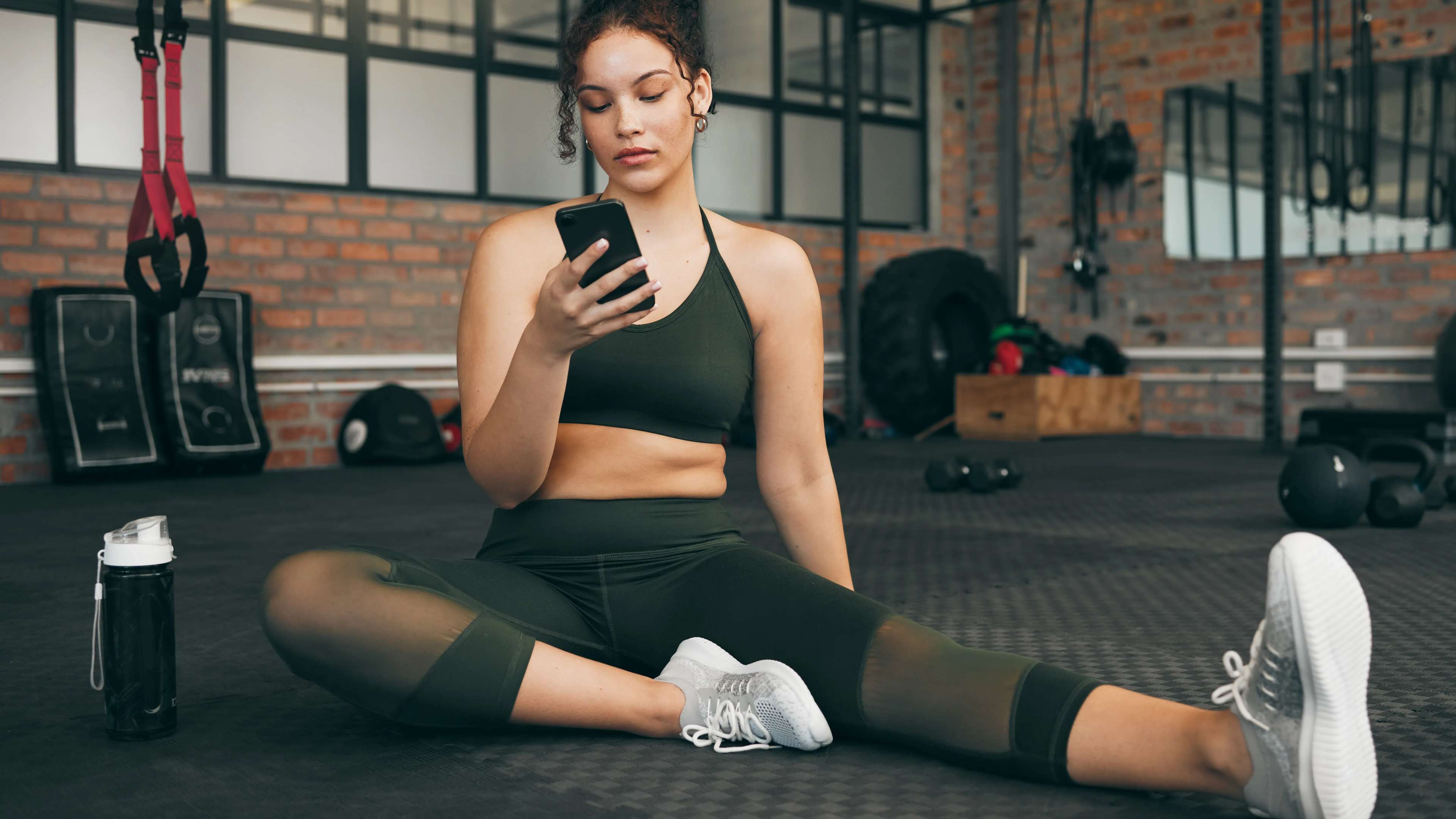 Eine Frau sitzt auf dem Boden im Fitnessstudio und schaut in ihr Smartphone