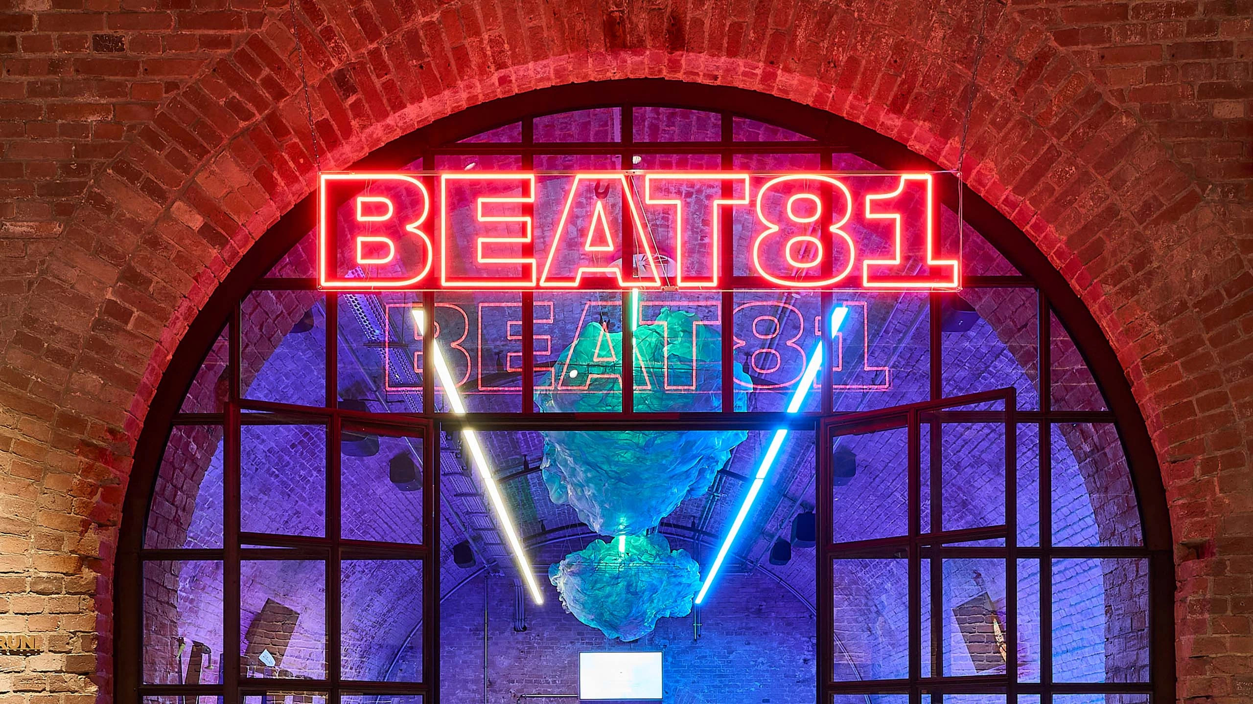 Eine große Glastür mit schwarzen Rahmen in einem Rundbogen einer roten Backsteinwand. Oben an der Tür hängt eine rotleuchtende Neonreklame mit dem Schriftzug BEAT 81. Der Schriftzug spiegelt sich in der Tür wider. An der Decke in dem Raum hinter der Glastür hängen helle Neonröhren, in der Mitte sind über der Neonröhre weiße Dekoelemente aufgehangen, grünlich schimmernd vom Licht. Mittig steht auf dem Bild der Schriftzug: Beat81 X JR Bötzow. Darunter ein klickbarer Button mit der Aufschrift Register now.