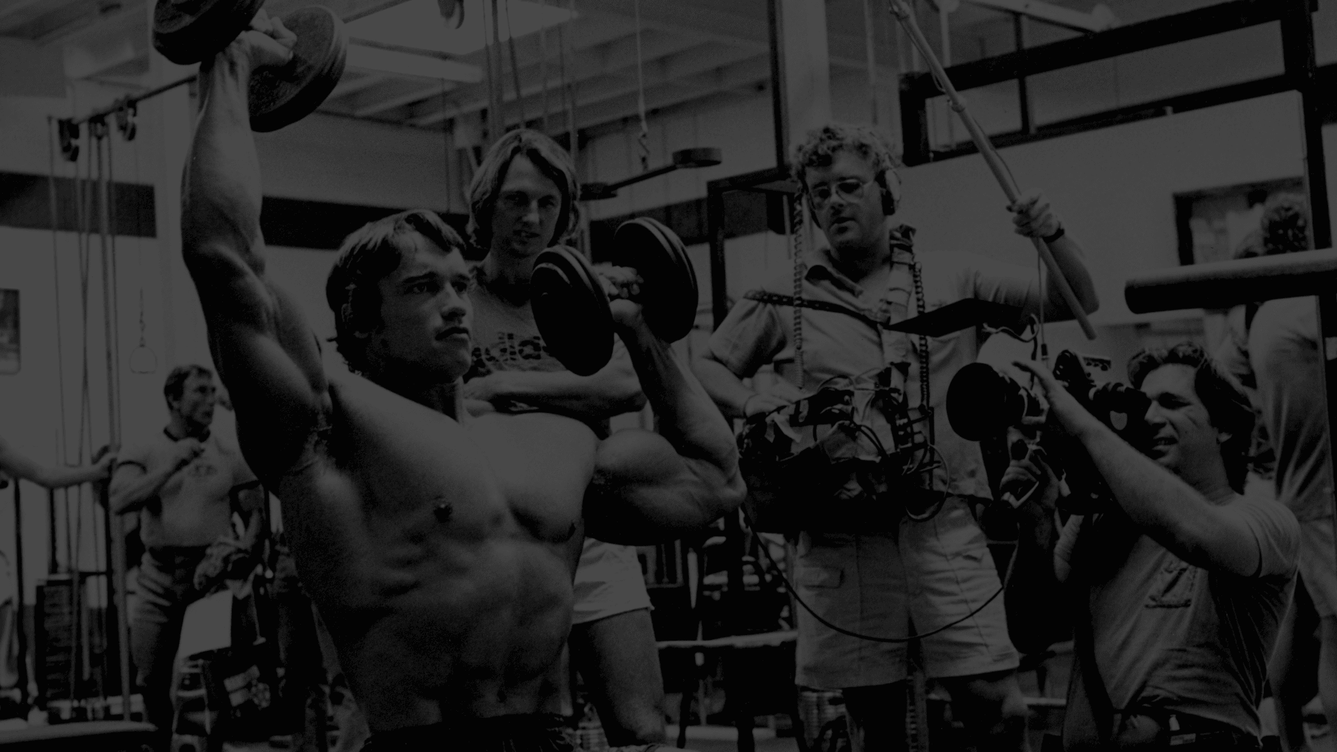Ein Kamerateam filmt Arnold Schwarzenegger beim Training mit Kurzhanteln im Gold's Gym Fitnessstudio