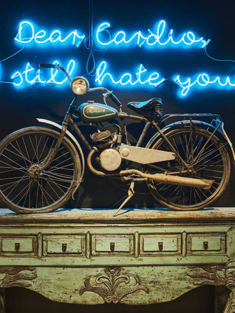 Ein altes Fahrrad mit Motor, steht auf einer antiken Kommode. Darüber ist eine leuchtende Neonreklame mit dem Schriftzug: Dear Cardio, still hate you.